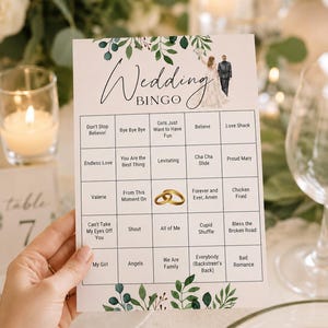 May include: A white rectangular Wedding Bingo game card featuring a green leaf border at the top and bottom, a small illustration of a couple, and a grid containing song titles and a central image of two gold wedding rings in black text.