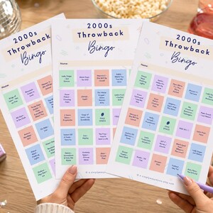 May include: Three sheets of 2000s Throwback Bingo cards with colorful squares and song titles. The cards are held by hands, with a purple pen resting on one. A pink flip phone and disco ball are in the background.