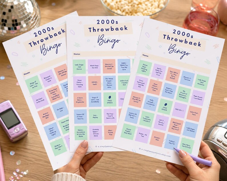 May include: Three sheets of 2000s Throwback Bingo cards with colorful squares and song titles. The cards are held by hands, with a purple pen resting on one. A pink flip phone and disco ball are in the background.
