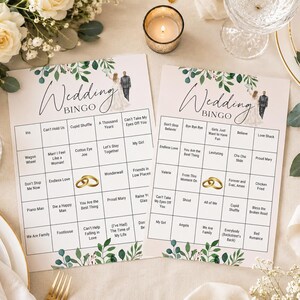 May include: Two white rectangular Wedding Bingo game cards featuring a green leaf border at the top and bottom. Each card displays a grid with song titles, a small illustration of a bride and groom, and two gold wedding rings in the center.