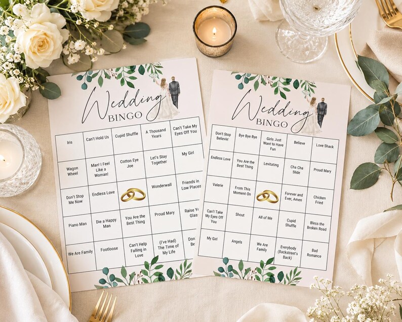 May include: Two white rectangular Wedding Bingo game cards featuring a green leaf border at the top and bottom. Each card displays a grid with song titles, a small illustration of a bride and groom, and two gold wedding rings in the center.
