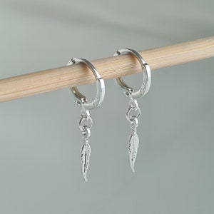 Könnte beinhalten: Silberne Creolen mit einem kleinen Feder-Anhänger, der an jeder Creole baumelt.