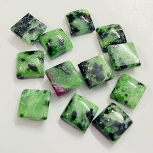 Puede incluir: Una colección de piedras preciosas de corte cuadrado. Las piedras son de un verde vibrante con inclusiones negras y rojas. Las piedras preciosas están pulidas y tienen bordes redondeados. Las piedras están dispuestas sobre una superficie blanca.