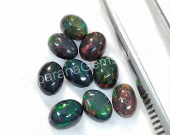 Puede incluir: Un conjunto de ópalos negros ovalados con destellos vibrantes de verde, rojo y dorado. Las piedras preciosas se exhiben sobre una superficie blanca, con unas pinzas plateadas en el fondo. Los ópalos tienen un acabado pulido y brillante.