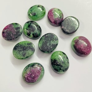May include: A collection of oval-shaped zoisite gemstones with a mix of green and magenta colors. The polished stones have a glossy finish and are arranged on a white surface. These gemstones are suitable for jewelry making.