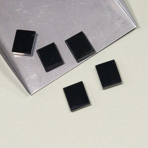 May include: Five small, rectangular, black objects are scattered on a silver tray and a light-colored surface. The objects appear to be polished and have a glossy finish. The arrangement suggests they may be components or decorative elements.