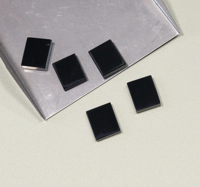 May include: Five small, rectangular, black objects are scattered on a silver tray and a light-colored surface. The objects appear to be polished and have a glossy finish. The arrangement suggests they may be components or decorative elements.