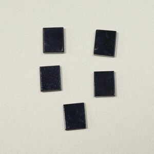May include: Five small, square, dark blue gemstones are arranged on a light background. Each stone has a polished, reflective surface, and they are slightly different in orientation. The gemstones appear to be of uniform size and shape.