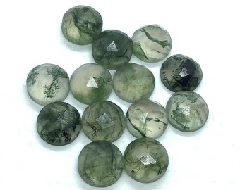 May include: A collection of faceted, round moss agate gemstones. The stones display a mix of translucent white and green hues, with intricate, moss-like inclusions. The gemstones are scattered against a white background, showcasing their natural beauty.