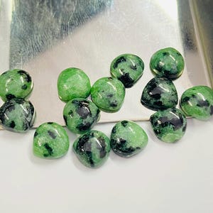 May include: A selection of polished green and black zoisite gemstones. The stones are oval and rounded, with a mix of light and dark green hues and black inclusions. The gemstones are displayed on a reflective surface.