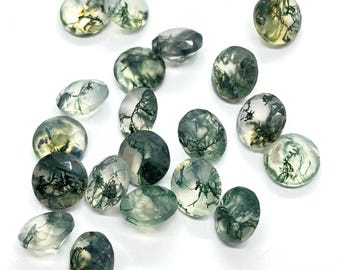 May include: A collection of moss agate gemstones in various shapes, including round and oval. The translucent stones feature green, tree-like inclusions, creating a natural, organic appearance. The gemstones are scattered on a white surface.