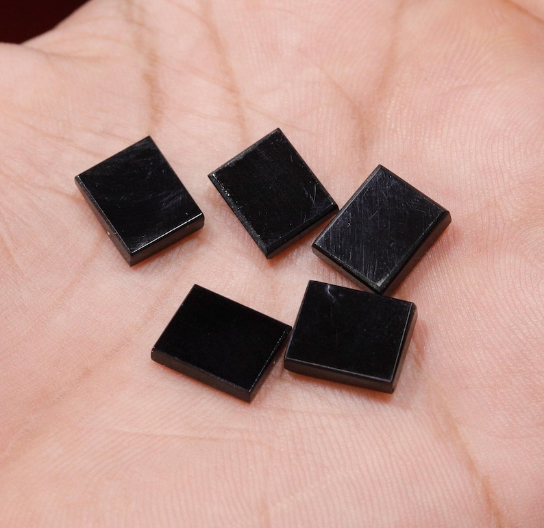 May include: Five small, rectangular, polished black gemstones are displayed. The stones are evenly sized and have a smooth, reflective surface. The gemstones are likely used for jewelry making or other crafts.