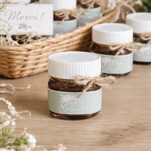 Peut inclure: Petits pots en verre remplis d'une substance foncée, probablement une confiture, avec des couvercles blancs et attachés avec de la ficelle. Chaque pot a une étiquette vert clair avec des détails floraux. Un panier tressé contient plusieurs pots, et une carte dit "Merci!"