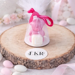 Peut inclure: Un savon rose en forme d'ours en peluche dans un sac en filet blanc, noué avec un ruban rose, présenté sur une tranche de bois. Un carré blanc avec une lettre est devant l'ours. Le logo "EKR" est sur un autocollant blanc.