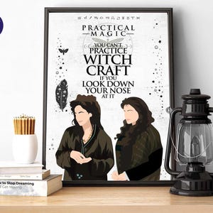 May include: A framed art print featuring two women and the text "PRACTICAL MAGIC - YOU CAN'T PRACTICE WITCH CRAFT IF YOU LOOK DOWN YOUR NOSE AT IT." A black lantern and a white vase with pencils are on a wooden surface.
