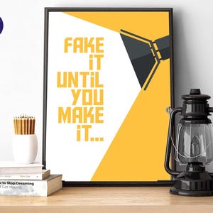 Könnte beinhalten: Ein gerahmter Druck mit dem Text "FAKE IT UNTIL YOU MAKE IT" in Gelb vor einem weißen und gelben Hintergrund. Eine schwarze Laterne und eine weiße Vase mit Bleistiften stehen auf einer Holzoberfläche.