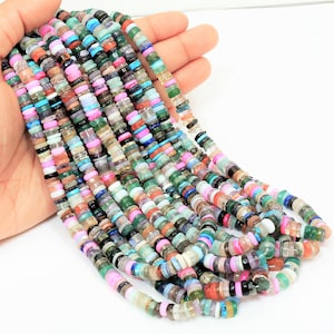 May include: A collection of multi-coloured gemstone beads, featuring pink, green, blue, and white hues. The beads are flat and disc-shaped, creating a layered effect. The beads are held in a hand against a white backdrop.
