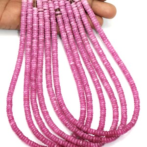 Op de afbeelding: Strengen roze edelsteen kralen. Elke streng bestaat uit kleine, ronde, platte kralen, aan elkaar geregen. De kralen hebben een consistente kleur en textuur, waardoor een uniforme uitstraling ontstaat. De kralen worden in een hand gehouden tegen een witte achtergrond.