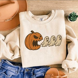 Puede incluir: Sudadera de color crema con un gráfico de Halloween. El diseño incluye una calabaza con una expresión amenazante y tres fantasmas de dibujos animados. La sudadera se combina con vaqueros azules y un sombrero marrón.