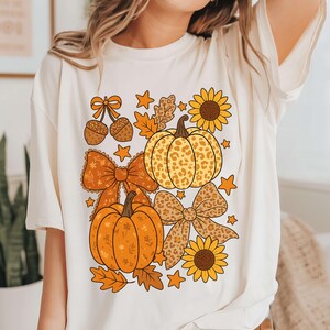 May include: Cream-coloured t-shirt with a fall-themed graphic. The design includes orange pumpkins, bows, sunflowers, acorns, and leaves. The pumpkins have leopard print and solid orange patterns. The shirt has a relaxed fit.