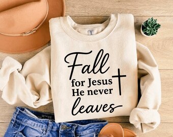 May include: Cream-colored sweatshirt with the text "Fall for Jesus He never leaves" in black script and a cross. The sweatshirt is paired with blue denim jeans, a brown hat, and sandals.