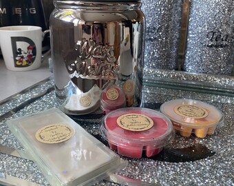 May include: A silver metal jar with the words "Wax Melts" on the front. The jar is sitting on a silver tray with three small wax melts in clear containers. The wax melts are red, orange, and white.
