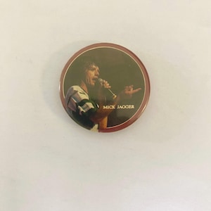 Könnte beinhalten: Ein runder Knopf mit rotem Rand und einem Bild von Mick Jagger, der in ein Mikrofon singt. Der Text "MICK JAGGER" ist unter dem Bild gedruckt.