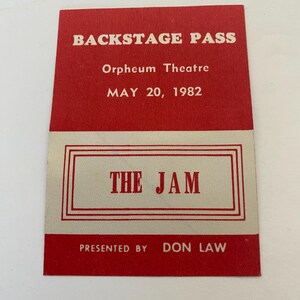 May include: A vintage backstage pass with a red and cream color scheme. The text "BACKSTAGE PASS" is at the top, followed by "Orpheum Theatre" and "MAY 20, 1982." The words "THE JAM" are in a rectangular box. "PRESENTED BY DON LAW" is at the bottom.
