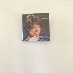 Może przedstawiać: Zbliżenie na vintage'owy guzik z fotografią Micka Jaggera. Guzik jest czarny z złotym napisem "Mick Jagger".