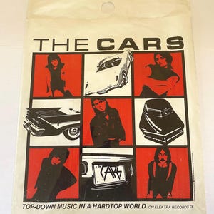 May include: A white and black promotional poster for the band The Cars. The poster features a grid of nine squares with images of the band members and cars. The text "THE CARS" is at the top of the poster. The text "TOP-DOWN MUSIC IN A HARDTOP WORLD ON ELEKTRA RECORDS" is at the bottom of the poster.