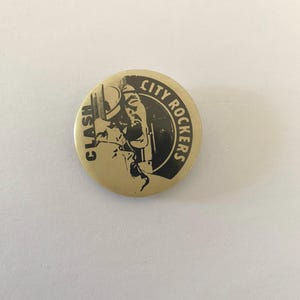 Może przedstawiać: Złoty guzik z czarno-białym obrazkiem osoby w kapeluszu trzymającej mikrofon. Wokół obrazka nadrukowany jest tekst "CITY ROCKERS" i "CLASH".