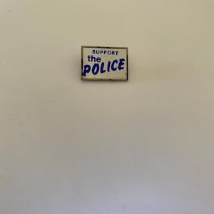 May include: A white enamel pin with blue lettering that reads "SUPPORT the POLICE".