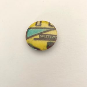 Peut inclure: Un bouton rond avec un motif jaune, turquoise et marron. Le texte "SPLIT ENZ" est imprimé au centre. Il s'agit d'un article vintage, probablement un badge.