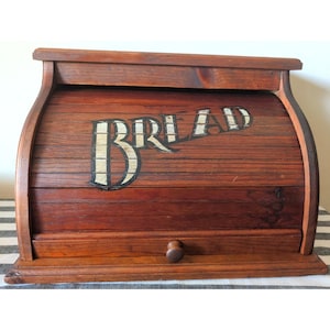 May include: A brown wooden bread box with a roll-top lid featuring the word BREAD painted in a distressed white font. The container has a rectangular base, a curved top section, and a small wooden knob on the front panel for opening the lid.