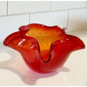 May include: A red and orange glass bowl with a ruffled edge and a textured surface. The bowl is made of bubble glass and has a unique, organic shape.
