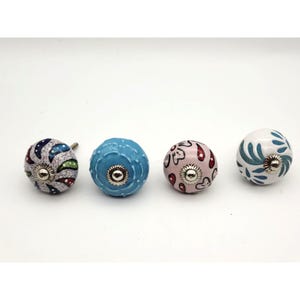 Peut inclure: Quatre boutons de placard ronds en céramique aux motifs distincts. Le premier est blanc avec des motifs cachemire bleus, verts et rouges. Le deuxième est bleu clair uni avec une texture en relief. Le troisième est rose clair avec des motifs floraux rouges et noirs. Le quatrième est blanc avec des motifs de feuilles bleues. Chaque bouton possède une pièce centrale en métal argenté.