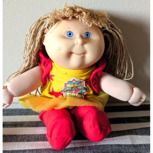 May include: A doll with light skin, blue eyes, and light brown yarn hair. The doll wears a yellow shirt with red stars and a graphic patch that reads Ballet, featuring red ruffled sleeves, a yellow tulle skirt, and solid red fabric trousers.
