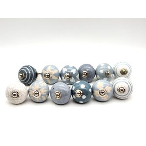May include: A collection of twelve decorative ceramic cabinet knobs. The knobs feature various designs, including stripes, stars, polka dots, and floral patterns, in shades of blue, gray, and white. Each knob has a silver-toned metal base.