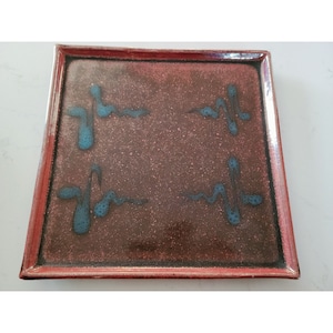May include: Square ceramic serving tray with a red-brown glaze and blue abstract designs. The tray has a raised rim.