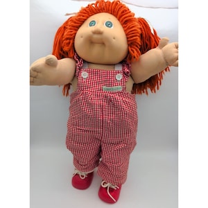 May include: A soft-sculpture doll with bright orange yarn hair, green eyes, and dimpled cheeks. The doll wears red and white checkered overalls with two white buttons at the chest and a small green label, paired with red shoes with white laces.