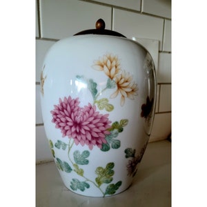 May include: White porcelain jar with a floral design. The jar has a lid with a wooden knob. The flowers are pink, yellow, and green.