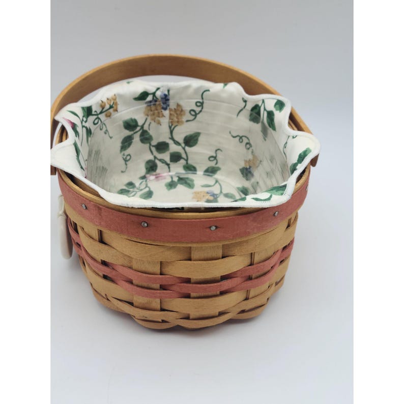 May include: A woven basket with a wooden handle and a pink band around the middle. The interior features a floral patterned fabric lining with green leaves and yellow and purple flowers. The basket is set against a white background.