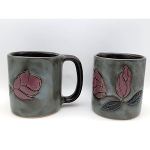 May include: Two ceramic mugs with a gray-blue glaze and a black handle. Each mug features a raised design of a pink flower with gold accents and green leaves. The interior of the mugs is black.