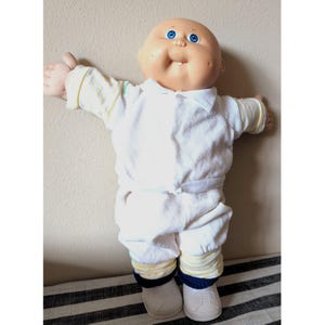 May include: A doll with a round, tan-coloured head, bright blue eyes, and a single visible tooth. The doll wears a white collared shirt with yellow striped sleeves, white trousers with yellow striped cuffs, dark blue socks, and white shoes.