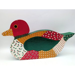 May include: A flat wooden duck cutout painted with a patchwork design. The head is red with a yellow beak, while the body features sections of green, red, and white patterns including holly leaves, berries, and small dots in a folk art style.