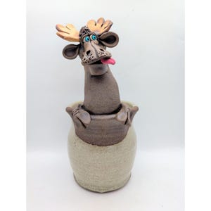May include: A ceramic sculpture of a brown moose with tan antlers, bright blue eyes, and a pink tongue sticking out. The moose is emerging from a light beige, rounded ceramic jar. The piece features a textured, earthy finish throughout.