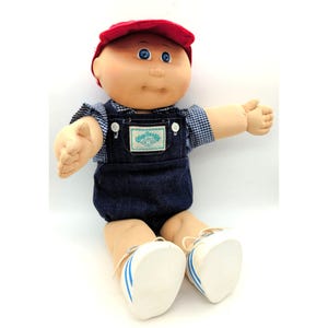 May include: A Cabbage Patch Kids doll with a round face, blue eyes, and yarn hair, wearing a red cap, a blue and white checkered shirt, and dark blue denim overalls with a white label on the chest. The doll wears white shoes with blue stripes.