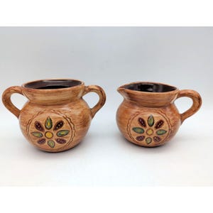May include: A ceramic sugar bowl with two handles and a matching creamer pitcher, both featuring a light brown glazed finish with a dark brown interior and a central hand-painted floral motif in yellow, green, and dark brown tones.