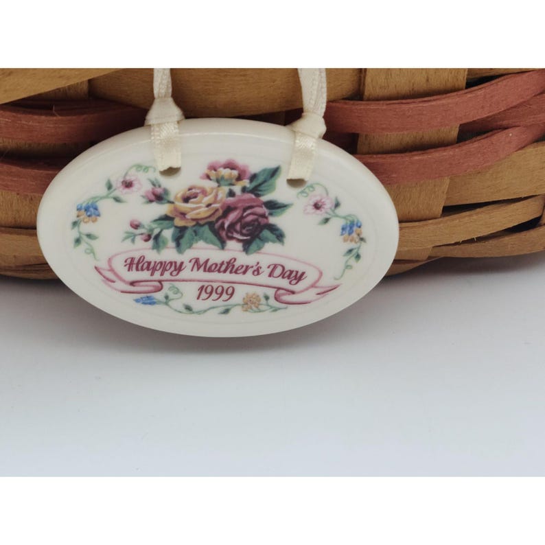 May include: An oval ceramic tag with the text "Happy Mother's Day 1999" and a floral design of roses and other flowers. The tag is attached to a woven basket with a light brown and reddish-brown pattern.