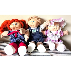 May include: Three dolls with yarn hair and soft bodies. The left doll has red pigtails and a blue plaid dress. The center doll has blonde hair and denim overalls. The right doll wears a floral dress with a matching bonnet featuring white lace trim.